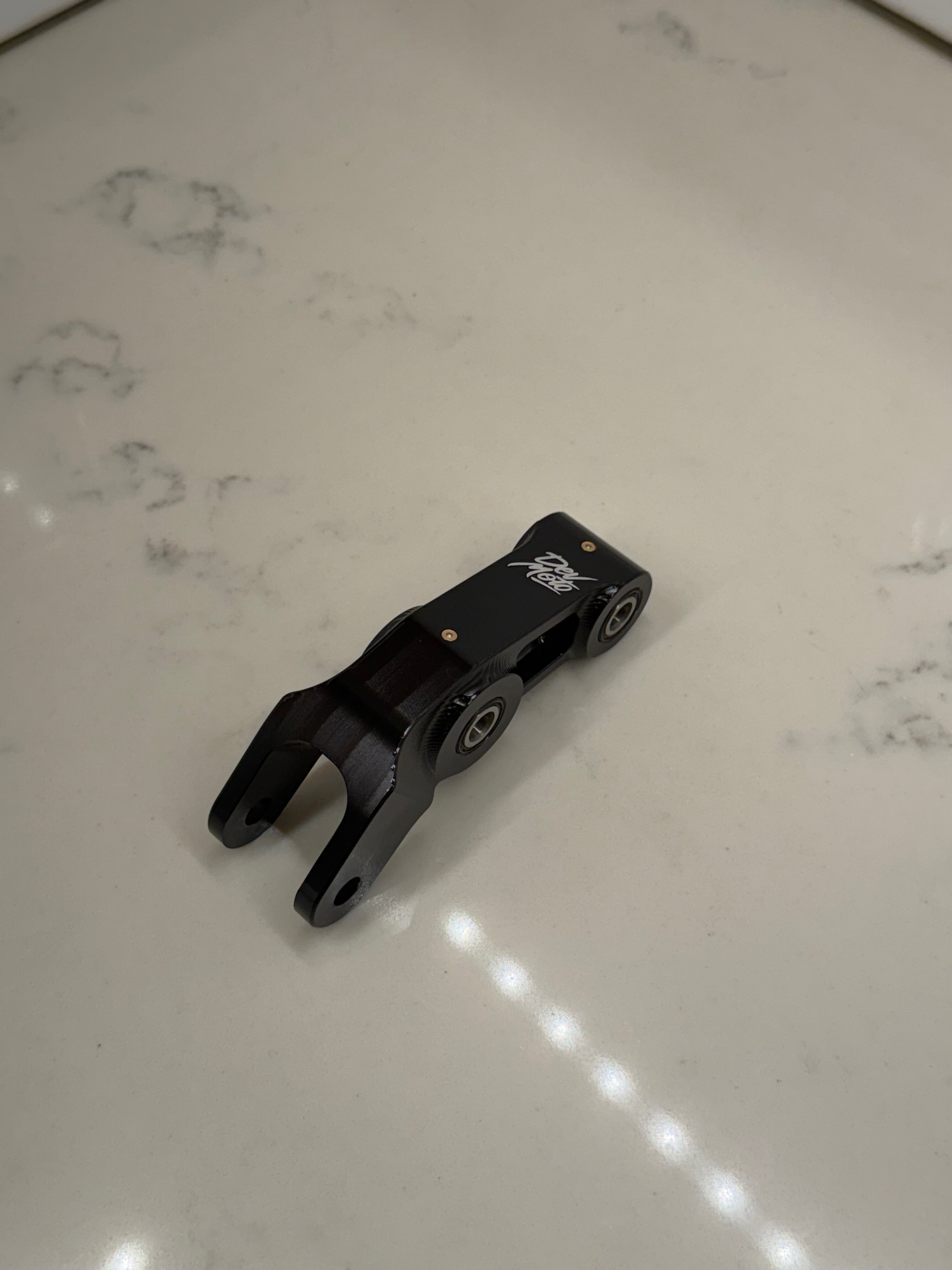 DevMoto E-Moto suspension linkage showing the metal linkage component with bolts on a neutral background.