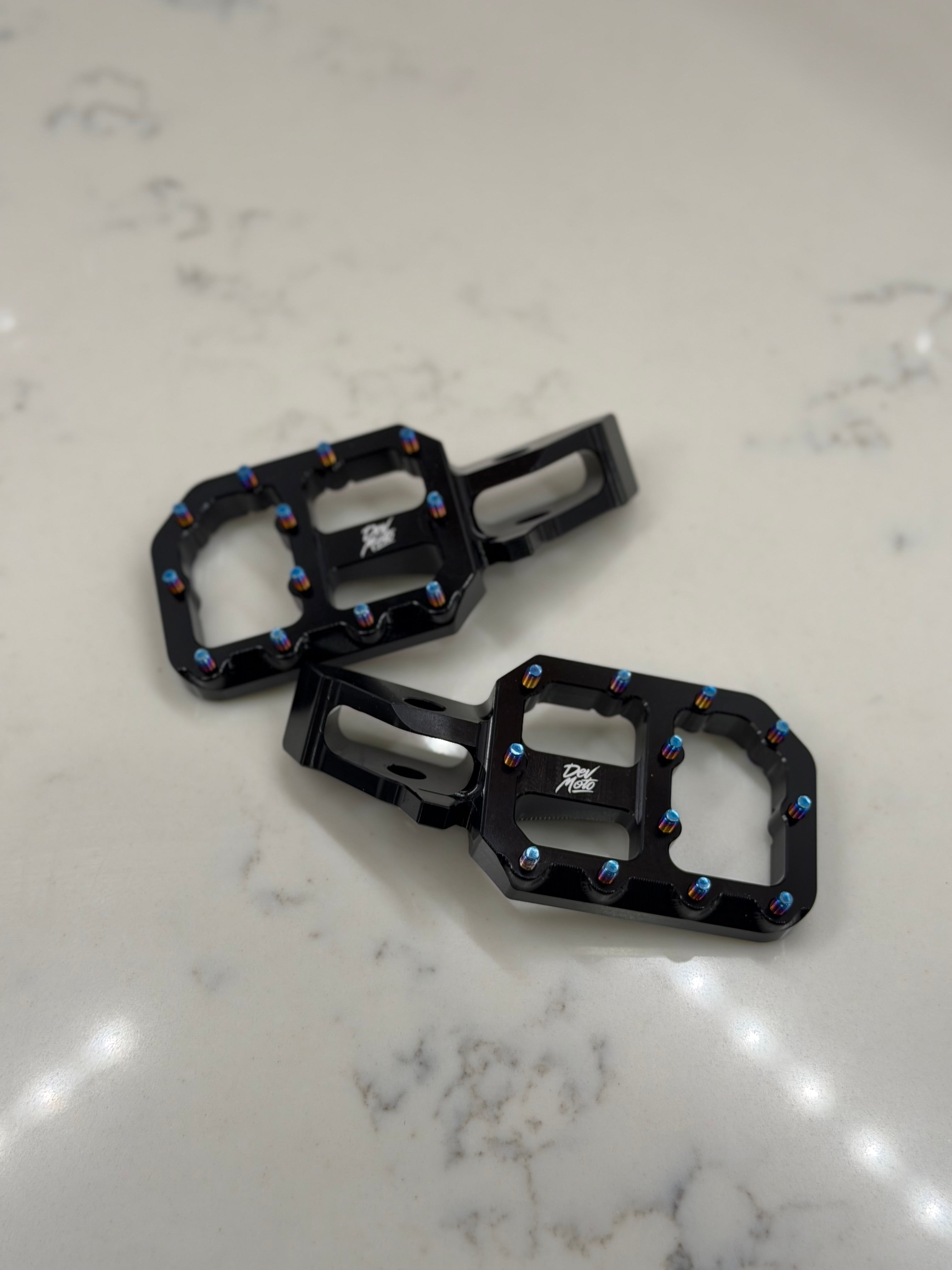 DevMoto E-Moto foot pegs for motorcycles, shown mounted on bike frame with textured metal surface and mounting hardware.